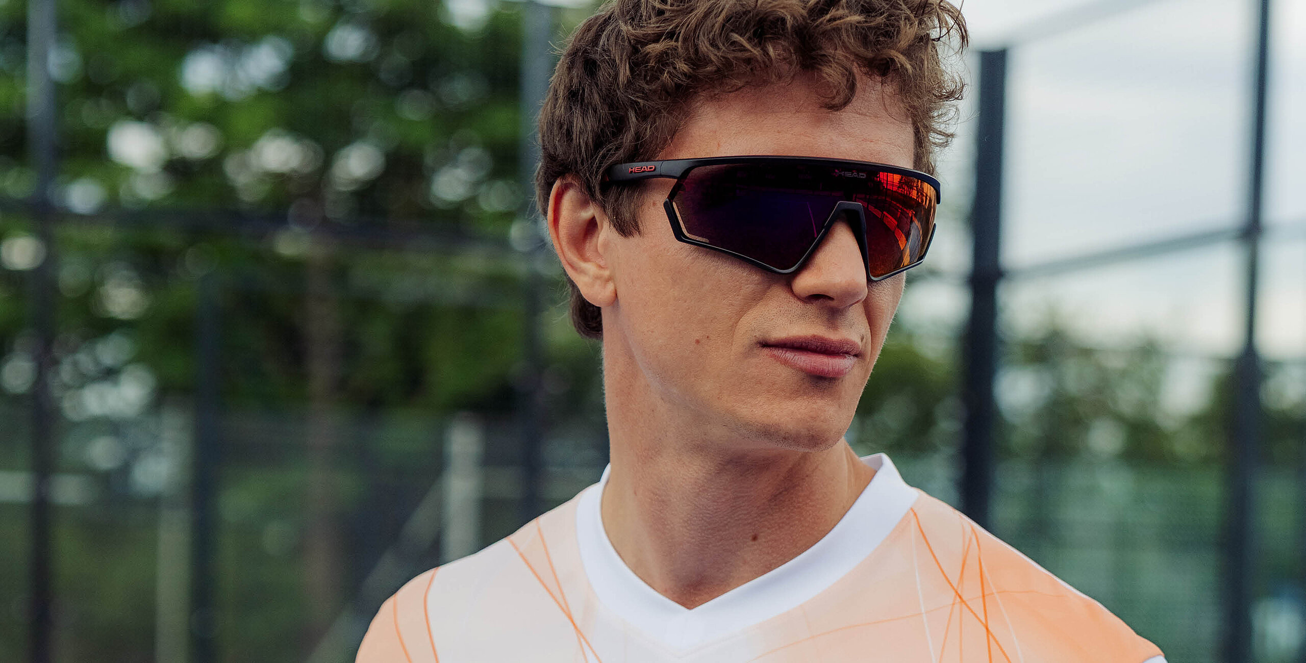 Close-up of a man on a padel court wearing black sport sunglasses with mirro lenses and HEAD red logo.