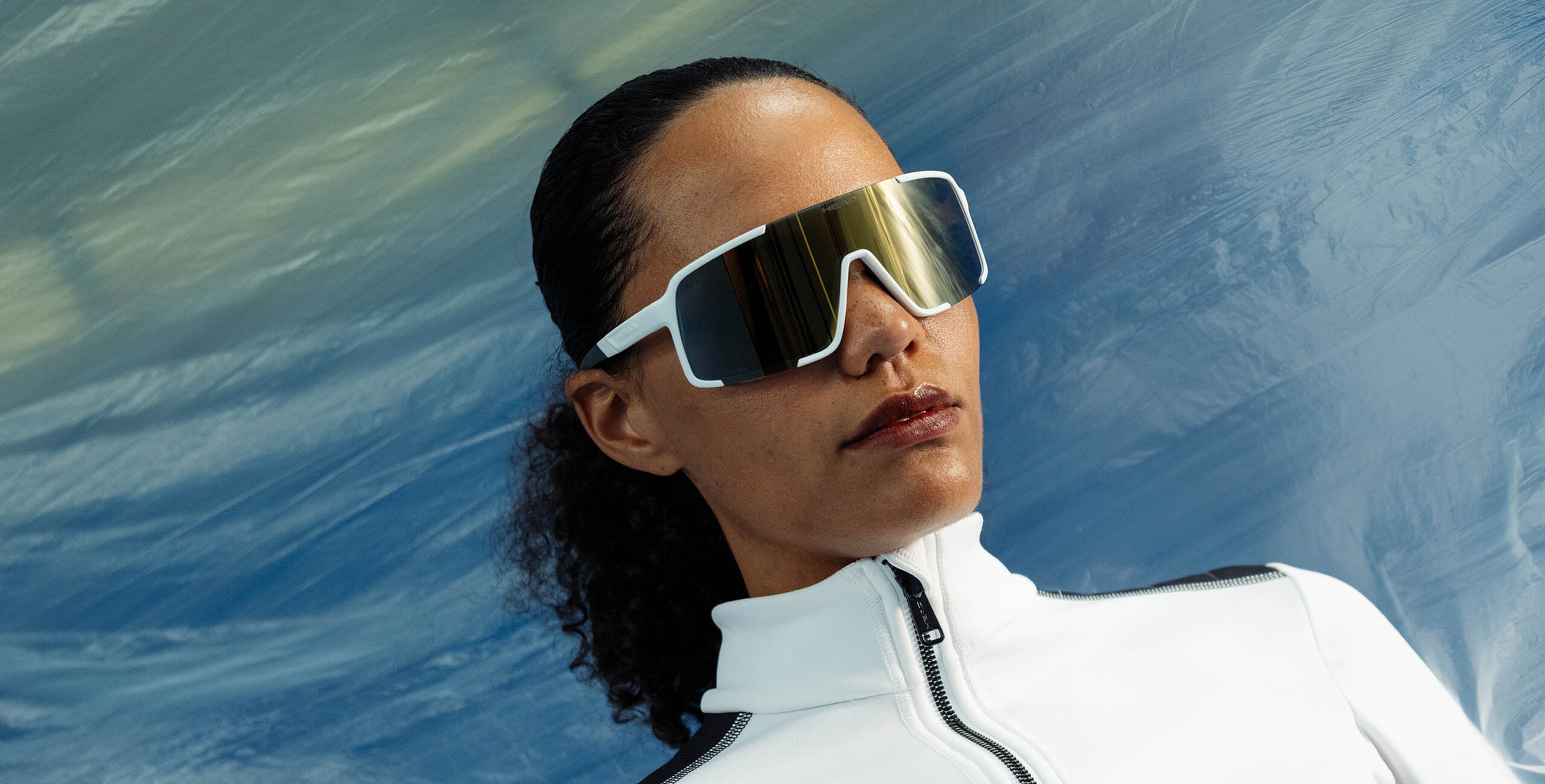 Woman wearing HEAD white sport sunglasses with gold mirrored lenses and a white zip-up jacket.