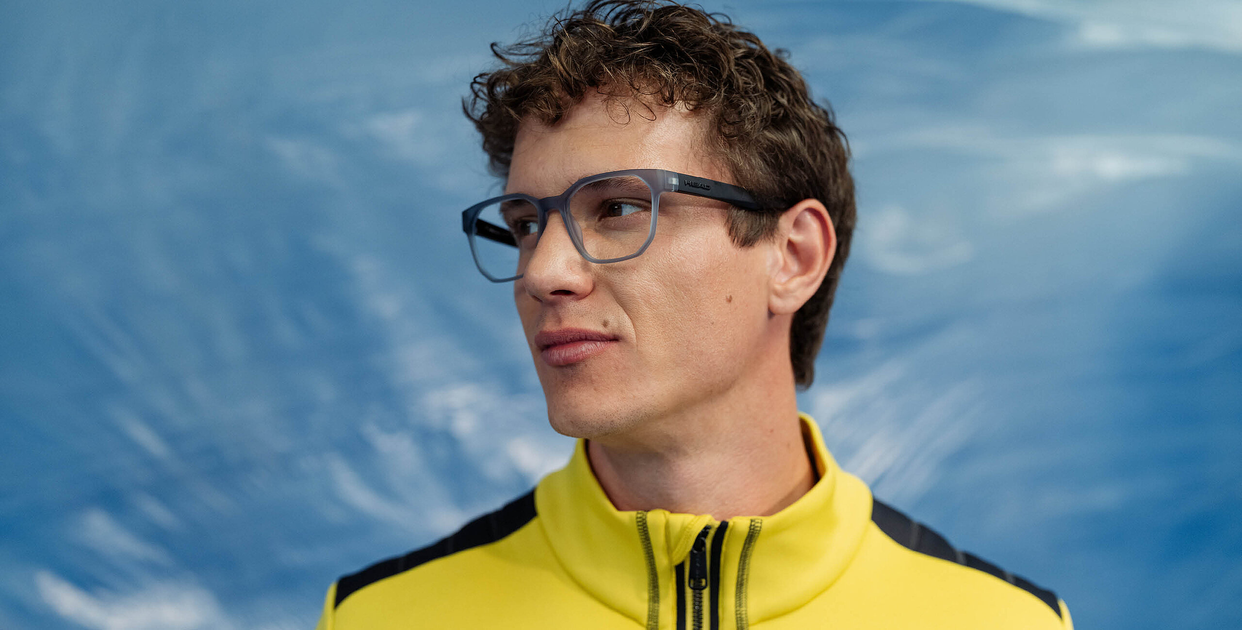 Man wearing HEAD blue glasses and a yellow and black jacket against a blurred blue sky background.