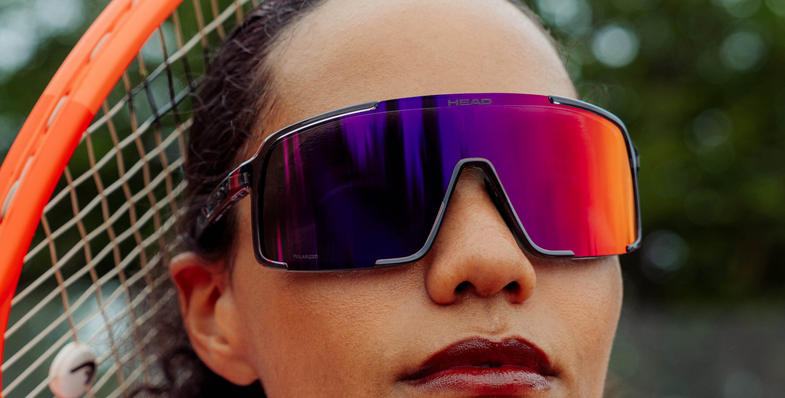 Close-up of a woman wearing HEAD shield sunglasses with a gradient of purple to orange mirror lens, with a tennis racket  visible on the left.