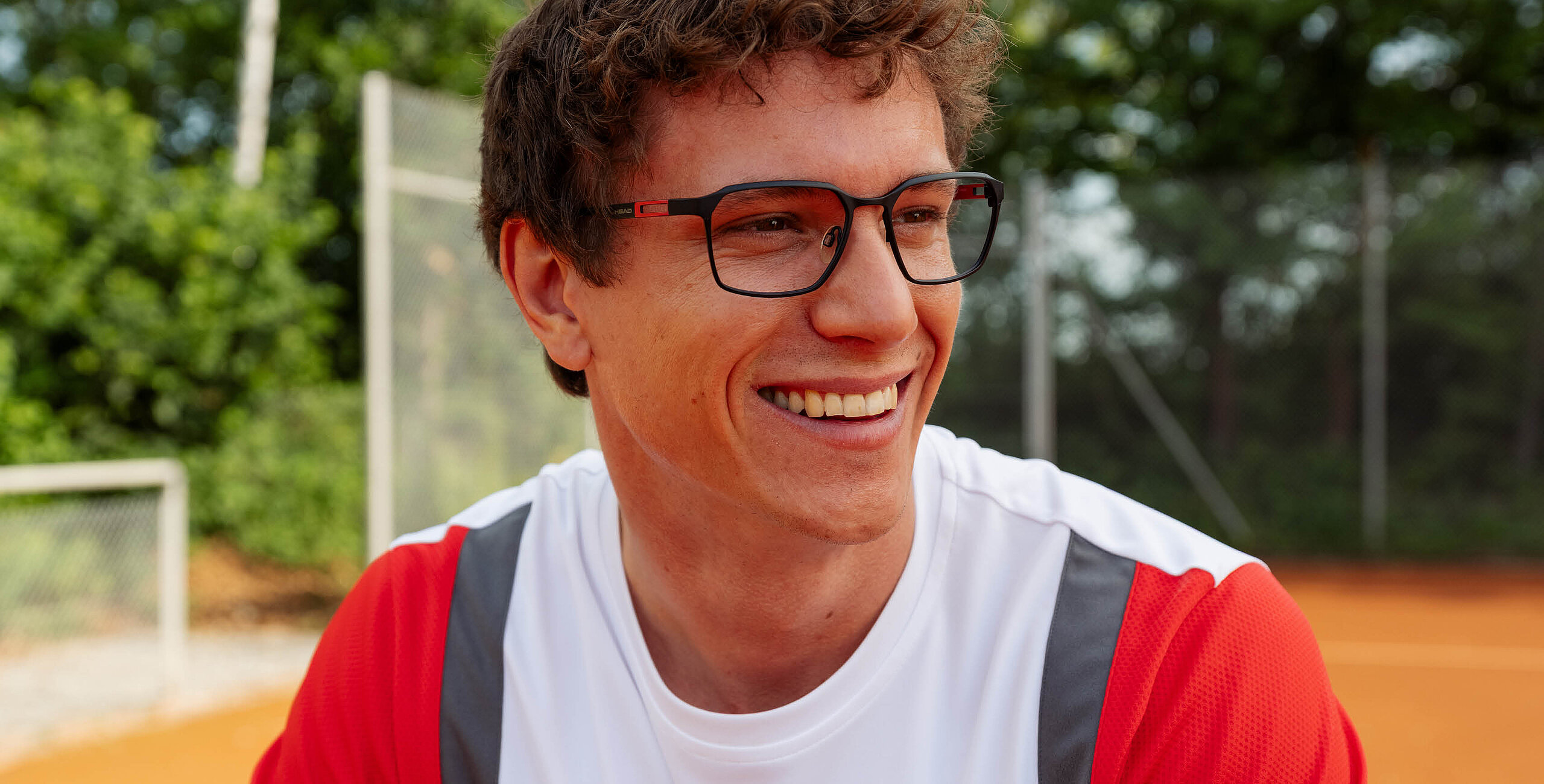Man wearing a HEAD black optical frame and a white and red shirt on a tennis court.